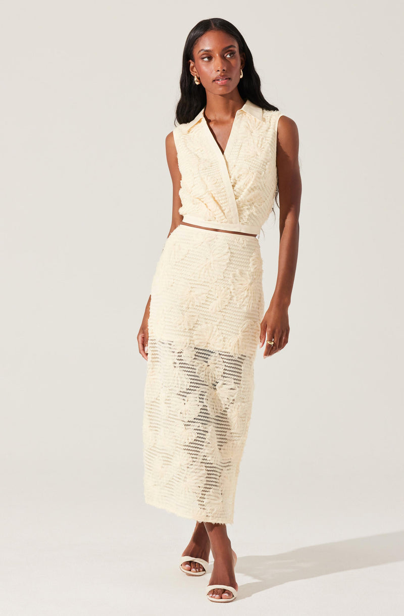 A woman is wearing a sleeveless, cream-colored knit two-piece outfit consisting of a V-neck crop top and a high-waisted, long skirt with slit and cut-out details. She is also wearing open-toe high heels.