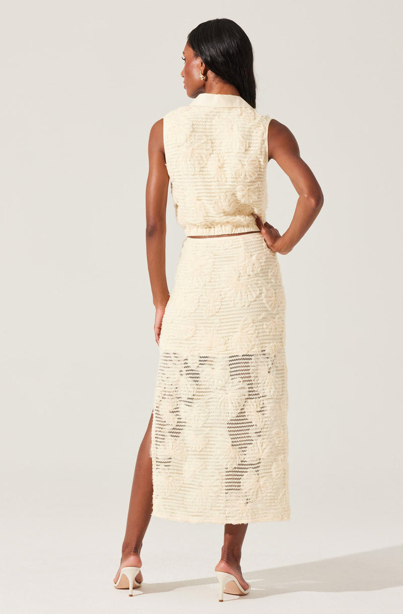 A woman is wearing a sleeveless, high-neck, cream-colored knit two-piece set consisting of a cropped top and a long skirt with a slit on one side. The knit fabric has a textured pattern with alternating open and closed stitches, creating a semi-sheer effect in some areas. She is also wearing nude high-heeled sandals.