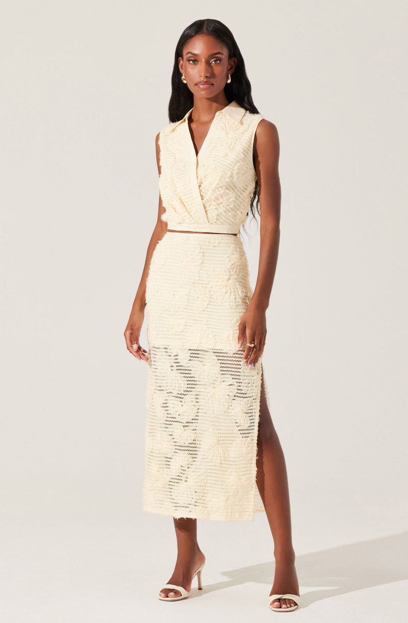 A woman is wearing a sleeveless, cream-colored outfit consisting of a textured, V-neck top with a collar and a matching high-waisted, ankle-length skirt with a slit on one side. She completes her look with open-toe high-heeled sandals.