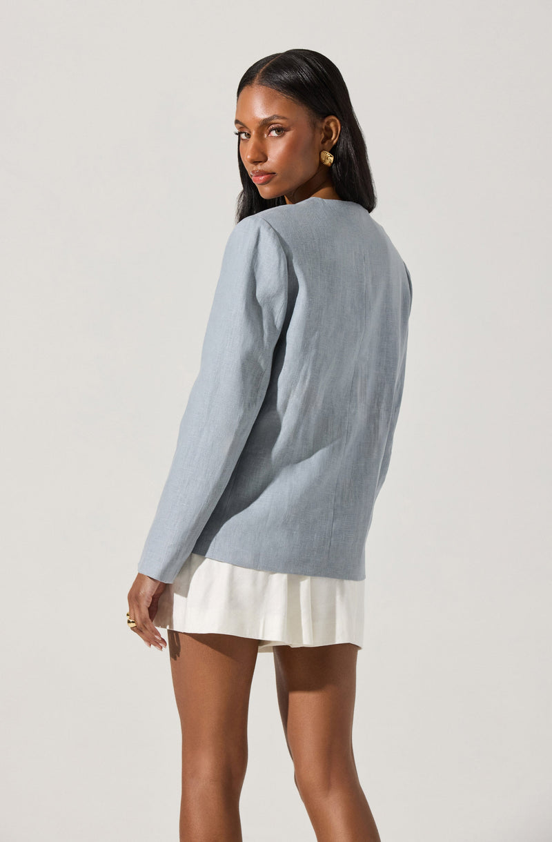 A woman with dark, straight hair is wearing a light gray, long-sleeved blazer-style top with minimal padding on the shoulders. She has paired it with a cream-colored, pleated mini skirt and is accessorized with gold earrings and a gold ring.