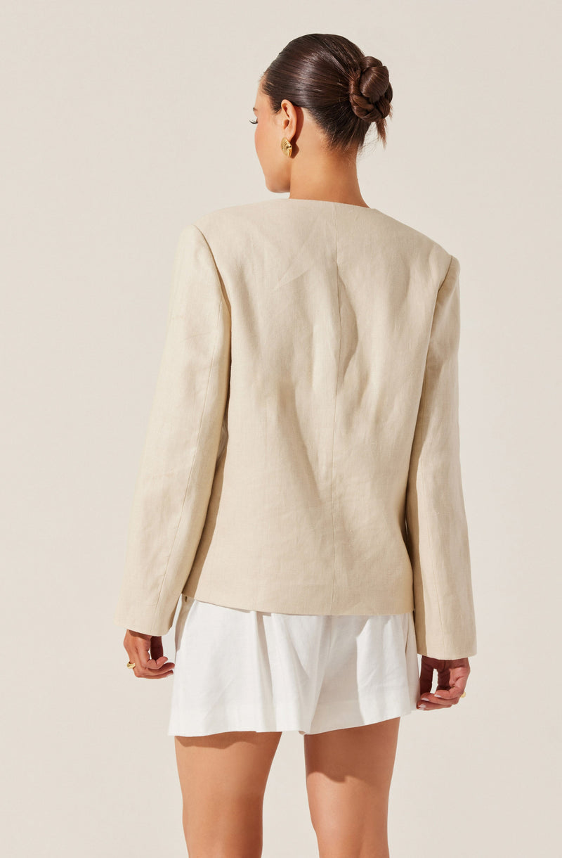 A woman is wearing a cream-colored blazer with structured shoulders and long sleeves. She pairs it with a white, pleated mini skirt. She has her hair styled in a neat, low bun and is accessorized with gold earrings.