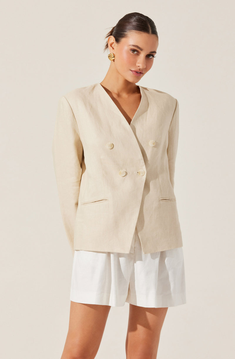A woman is wearing a light-colored, double-breasted blazer with large buttons and two small, horizontal slit pockets. She is also dressed in matching light-colored high-waisted shorts. The outfit has a neutral, beige tone, and she accessorizes with gold earrings.
