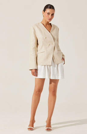 A woman is wearing a beige, double-breasted blazer with long sleeves and a V-shaped neckline. She pairs it with a white, pleated mini skirt and white high-heeled sandals.