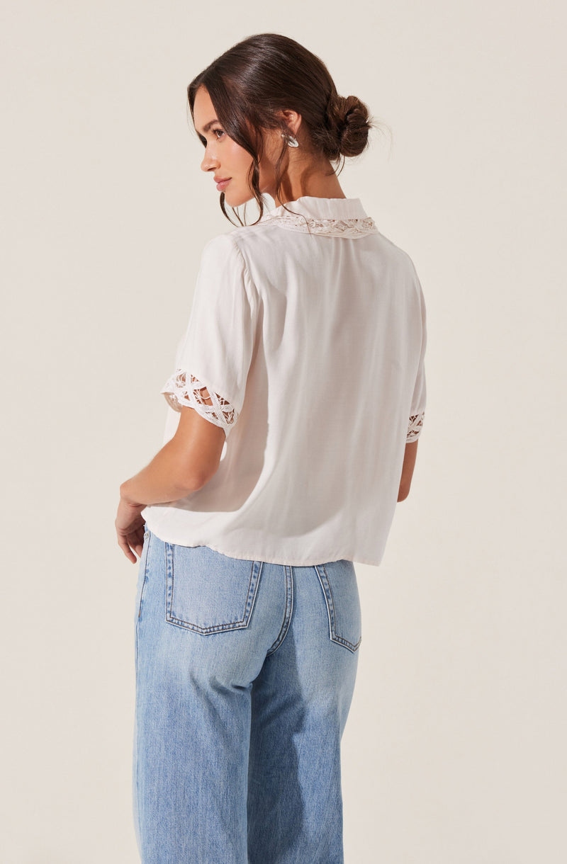 A woman is wearing a white short-sleeved blouse with lace trim on the sleeves and collar. She pairs it with light blue, high-waisted jeans. Her hair is styled in a low, loose bun.