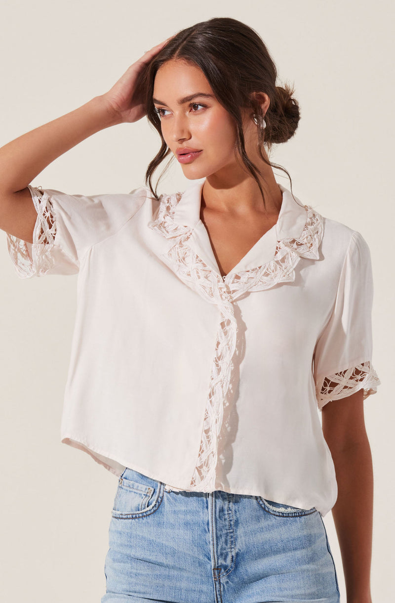 A woman is wearing a short-sleeved, cream-colored blouse with lace detailing on the collar, sleeves, and front placket. The blouse has an open collar and a slightly cropped, relaxed fit. She is also wearing light blue denim jeans.