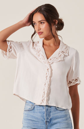 A woman is wearing a short-sleeved, cream-colored blouse with lace detailing on the collar, sleeves, and front placket. The blouse has an open collar and a slightly cropped, relaxed fit. She is also wearing light blue denim jeans.