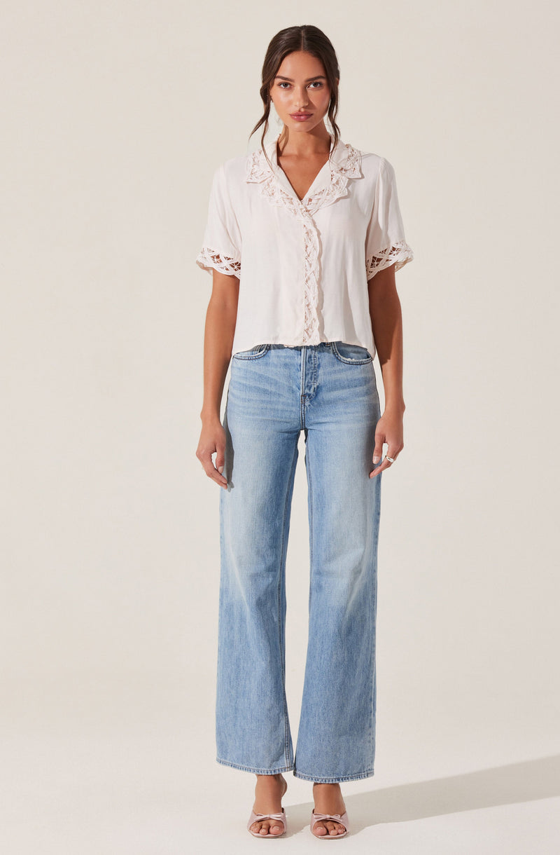 A woman is wearing a white short-sleeved blouse with lace trim details on the sleeves, collar, and front. She pairs it with light blue wide-leg jeans that reach the floor. She is also wearing open-toed, high-heeled sandals.