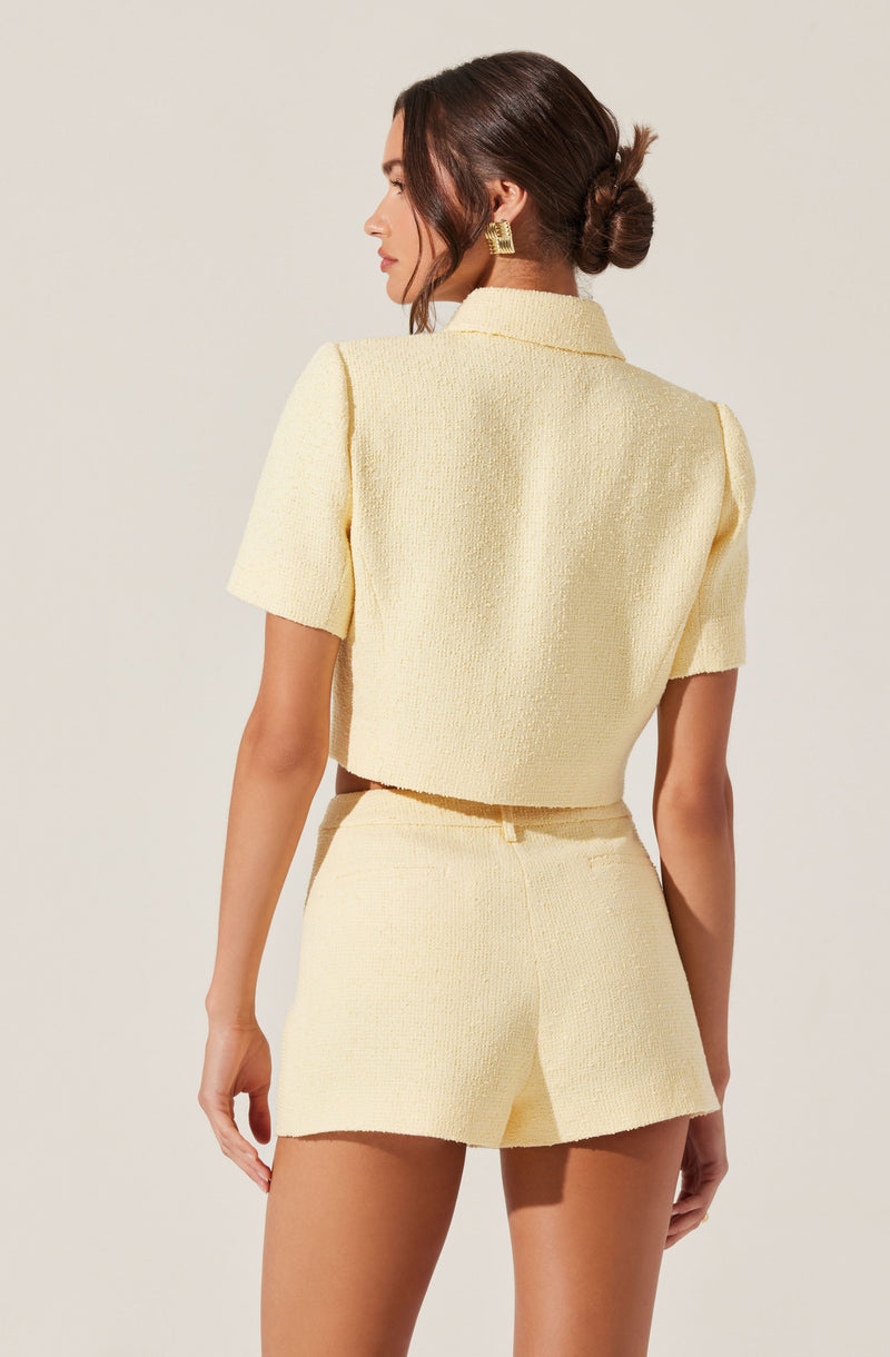 A woman is wearing a light yellow textured short-sleeve jacket with a collar, paired with matching high-waisted shorts. The outfit has a soft, woven appearance, and she accessorizes with large gold geometric earrings.