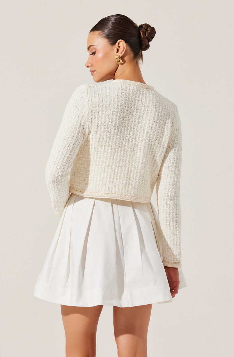 A woman is wearing a cream-colored textured knit sweater with long sleeves and a cropped fit, paired with a high-waisted, white pleated skirt. She has accessorized with large, gold hoop earrings. Her hair is styled in a sleek bun.