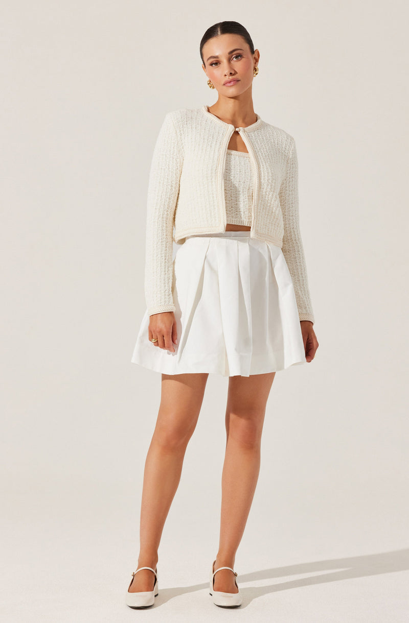 A woman is wearing a textured, cream-colored cardigan with a round neckline and an open front. Underneath, she has on a matching crop top. She pairs this with a high-waisted, white pleated skirt that falls above the knees. She completes the outfit with white high-heeled shoes with ankle straps.