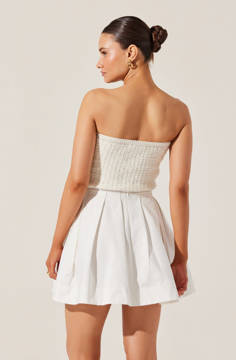 A woman is wearing a strapless, knitted, cream-colored top and a high-waisted white skirt with pleats.