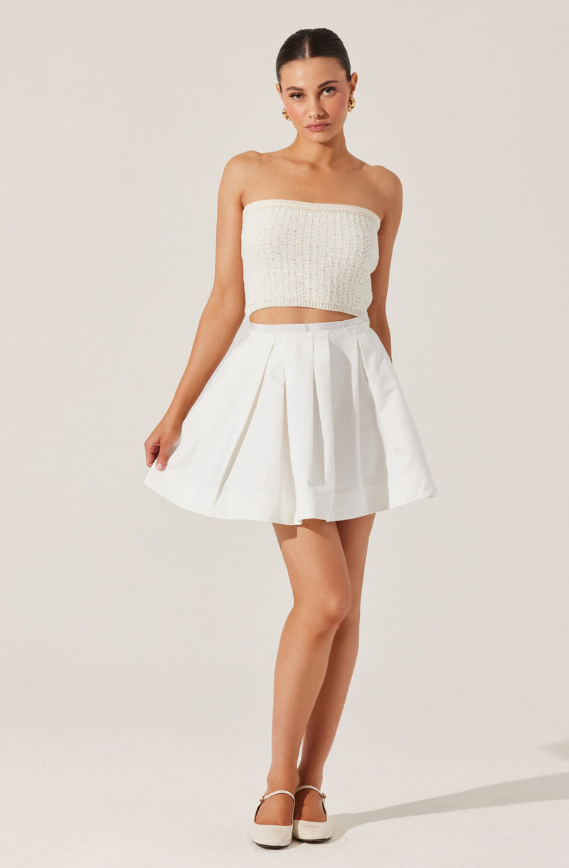 A woman is wearing a strapless, off-white, textured crop top and a high-waisted, white pleated skirt. She has on white heels with ankle straps.