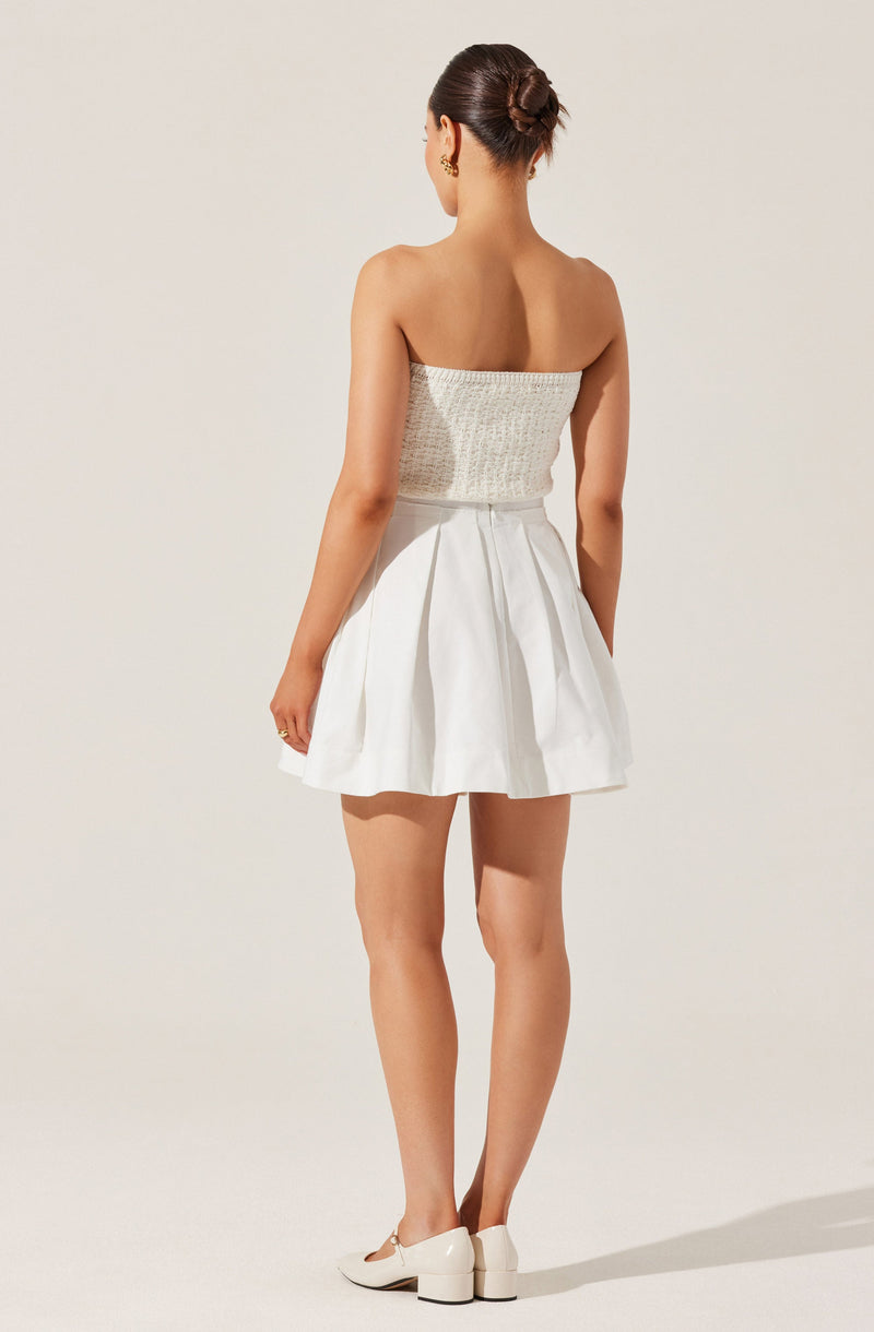 A woman is wearing a strapless top with a textured, knitted appearance in a light, neutral color. She has paired it with a high-waisted, white, pleated skirt that has a slightly flared silhouette. She is also wearing white heeled shoes. Her hair is styled in a neat bun.