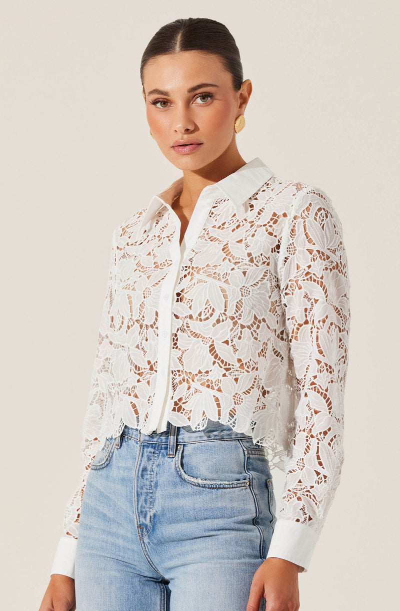 A woman is wearing a white, long-sleeved, button-up blouse made of lace with an intricate floral pattern. The blouse has a collar and scalloped edges at the hem and cuffs. She is also wearing light blue high-waisted jeans.