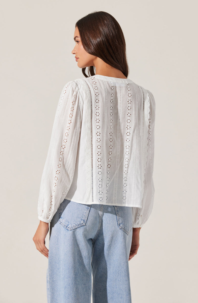 A woman is wearing a white long-sleeve blouse with a round neckline. The blouse features vertical rows of eyelet embroidery and cut-out patterns, giving it a delicate, textured appearance. The sleeves are slightly puffed and gathered at the cuffs. She is also wearing light-wash, loose-fitting jeans with visible stitching and back pockets.