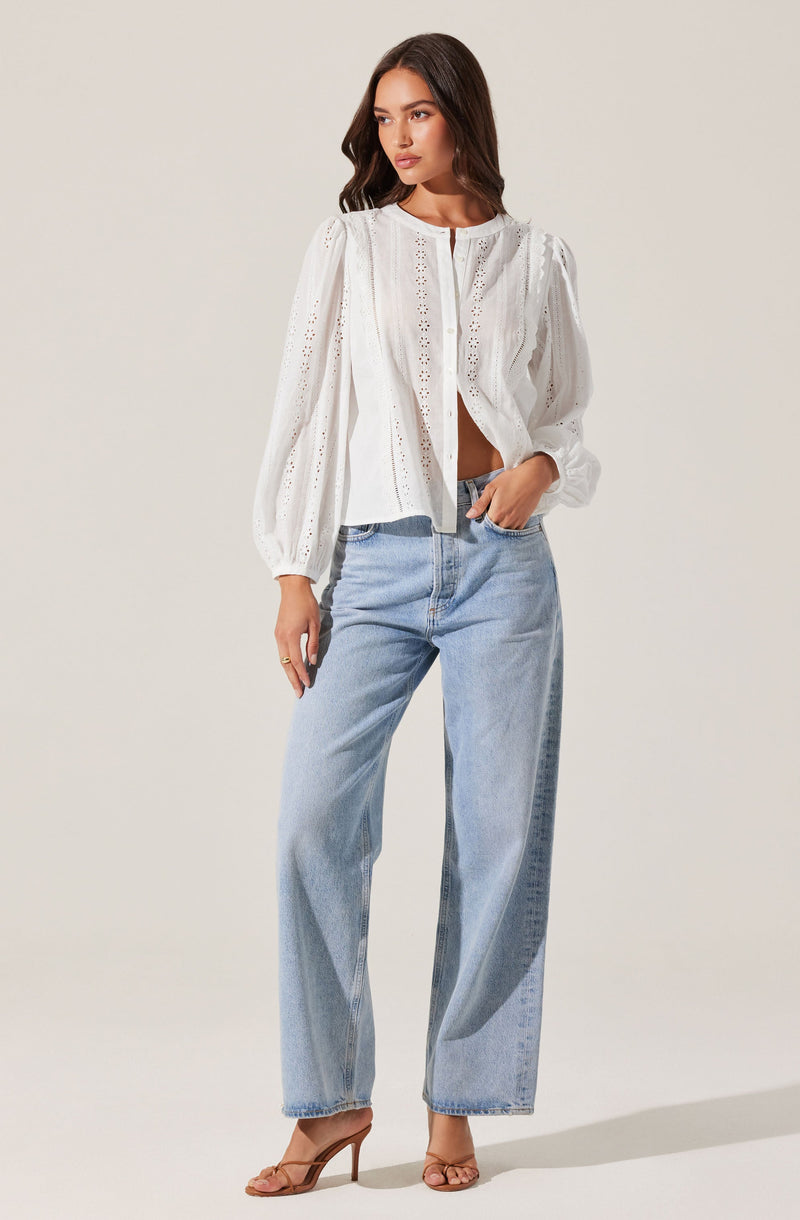A woman is wearing a white, long-sleeved blouse with eyelet embroidery details, puffed sleeves, and a loose fit. She paired it with high-waisted, light-wash, wide-leg jeans and beige high-heeled sandals.