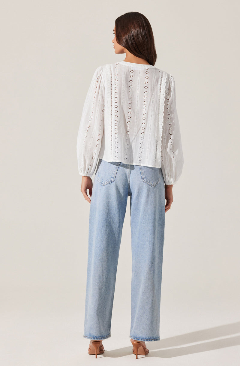 A woman is wearing a white, long-sleeved blouse with eyelet embroidery and a slightly loose fit. The blouse features vertical stripes of decorative cut-out details and has puffed sleeves that gather at the wrists. She pairs it with light-wash, high-waisted, straight-leg jeans and nude high-heeled shoes.