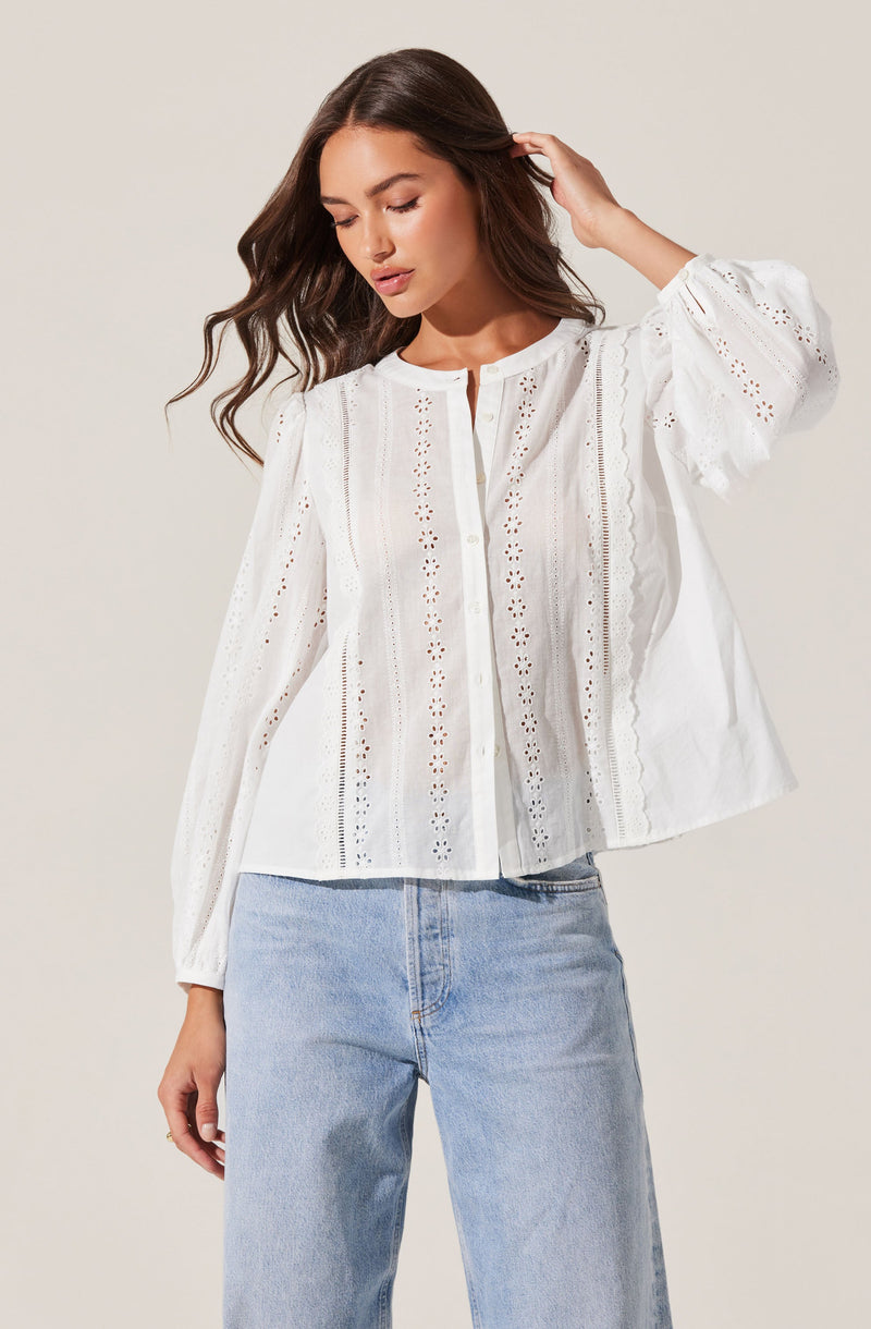 A woman is wearing a white, long-sleeved blouse with intricate eyelet embroidery and vertical lace details, featuring a round neckline and front button closure. She pairs it with light blue, high-waisted jeans.