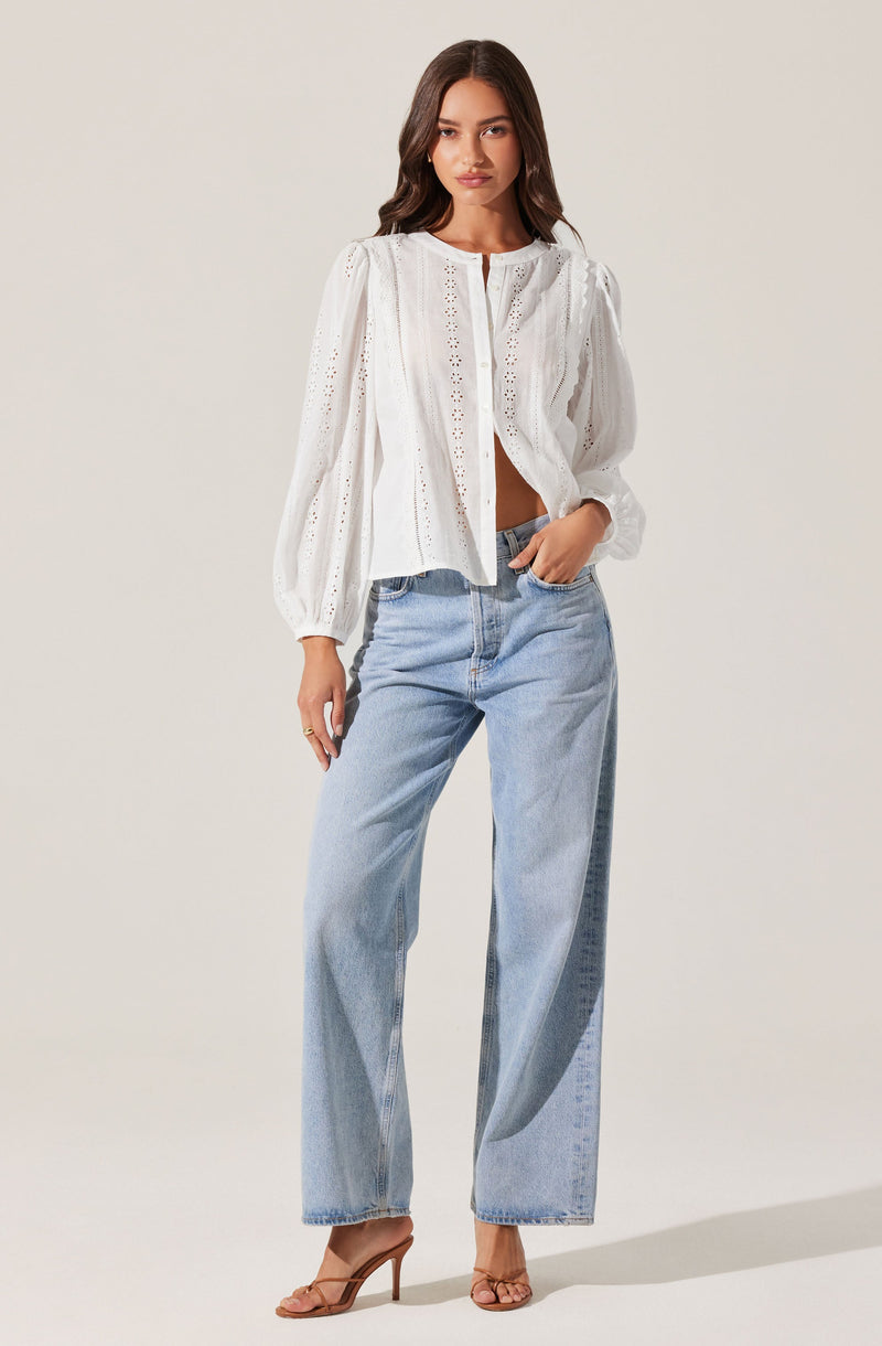 A woman is wearing a white, long-sleeved blouse with eyelet embroidery and a round neckline, paired with high-waisted, light-wash, wide-leg jeans. She is also wearing tan high-heeled sandals.