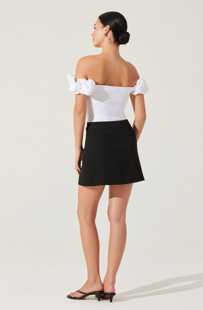 A woman is wearing an off-the-shoulder white top with puffed short sleeves and a fitted bodice, paired with a black mini skirt that has a subtle slit at the back and a small pocket on the right side. She is also wearing black open-toe heels with a small heel.