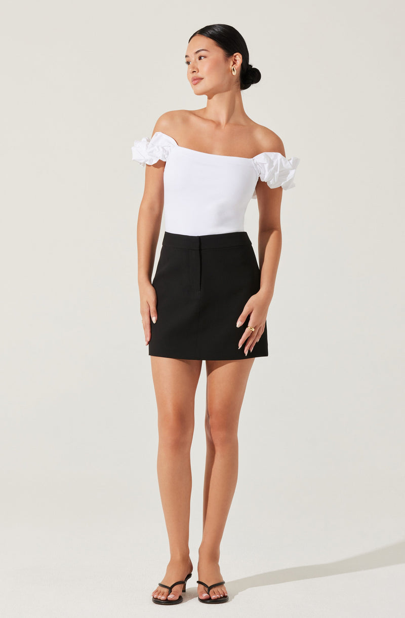 A woman wearing an off-the-shoulder white top with ruffled sleeves and a fitted bodice, paired with a high-waisted black mini skirt. She is also wearing black strappy high-heeled sandals and simple earrings.