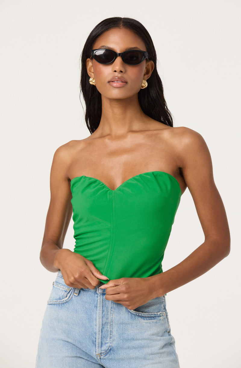 A woman is wearing a strapless, bright green top with ruching details, paired with light blue high-waisted jeans. She has on black sunglasses, gold hoop earrings, and has long, dark hair.