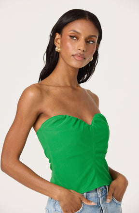 A woman is wearing a strapless, bright green top with a gathered detail along the bust and vertical seam down the front. She is also wearing light blue denim jeans and gold hoop earrings.