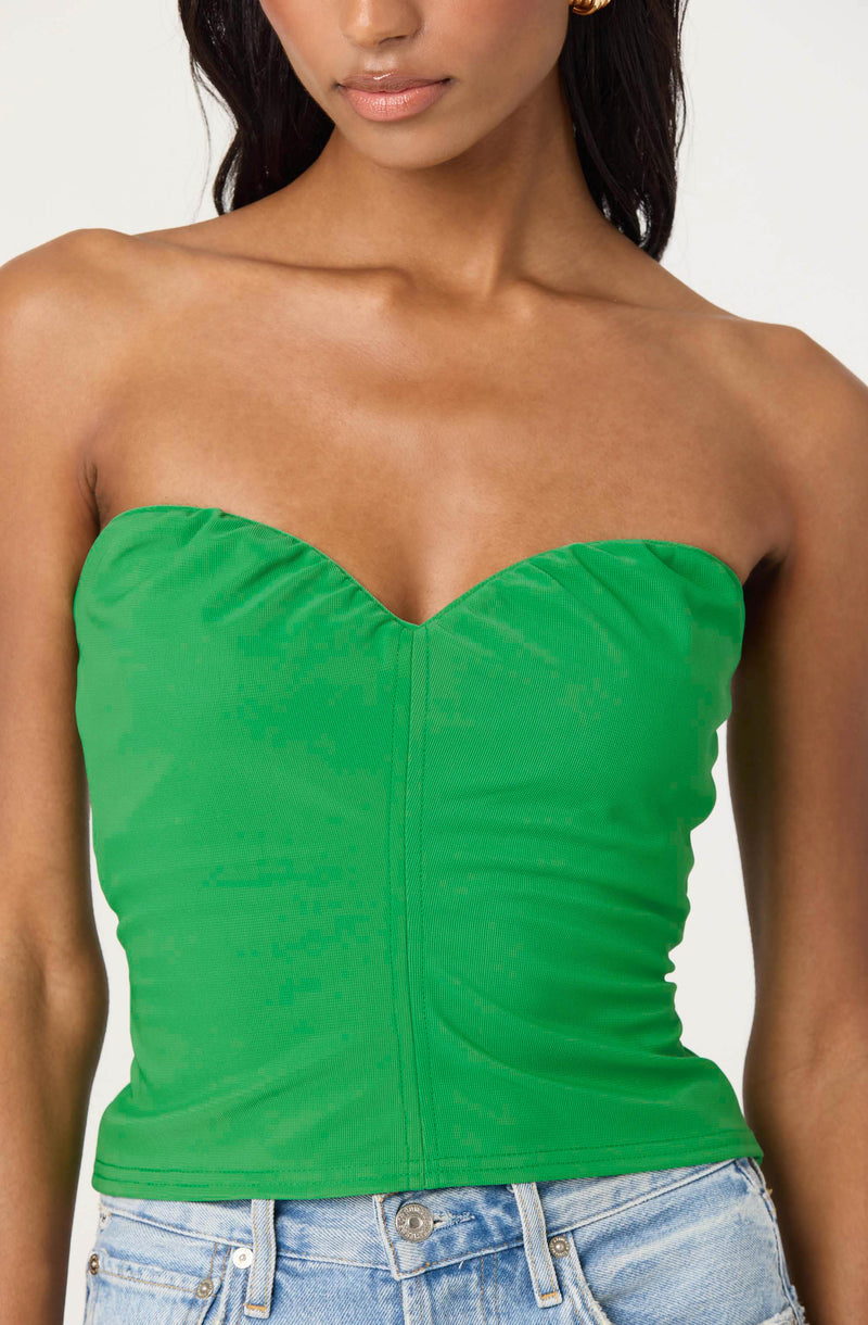 A woman is wearing a strapless, fitted green bustier top with a center seam detail. The top is smooth and slightly ruched on the sides. She is also wearing light-wash denim jeans.
