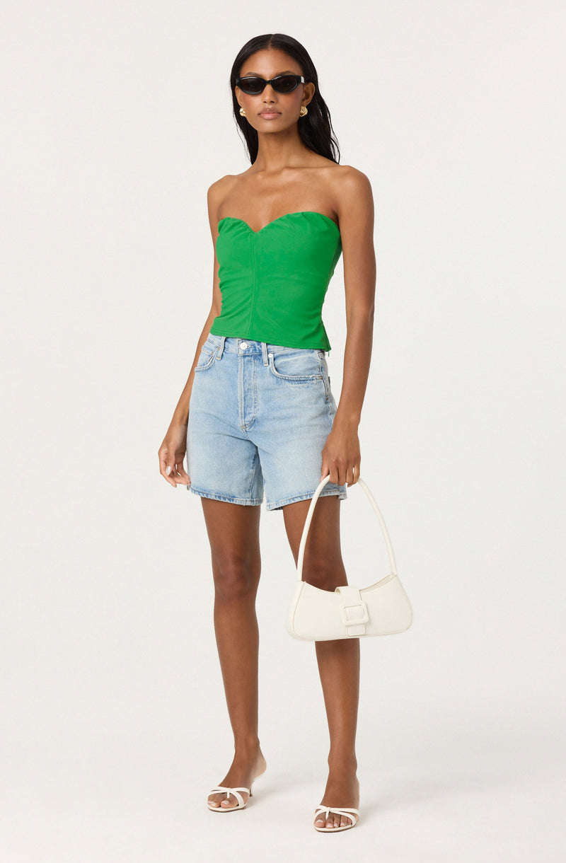 A woman is wearing a bright green strapless top with a ruffled neckline, paired with light blue high-waisted denim shorts. She has on black sunglasses, gold earrings, and white high-heeled sandals. She is carrying a small white handbag with a curved design and a buckle detail.