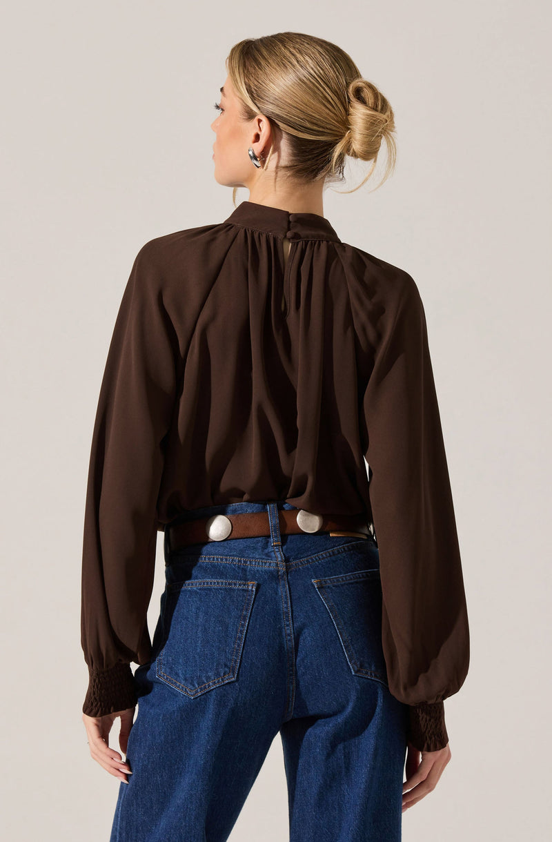 A woman is wearing a long-sleeved, dark brown blouse with gathered fabric at the back neckline and smocked cuffs. The blouse appears to be made of a silky or satin-like material. She pairs it with high-waisted blue jeans and a brown belt with white circular buckle details.
