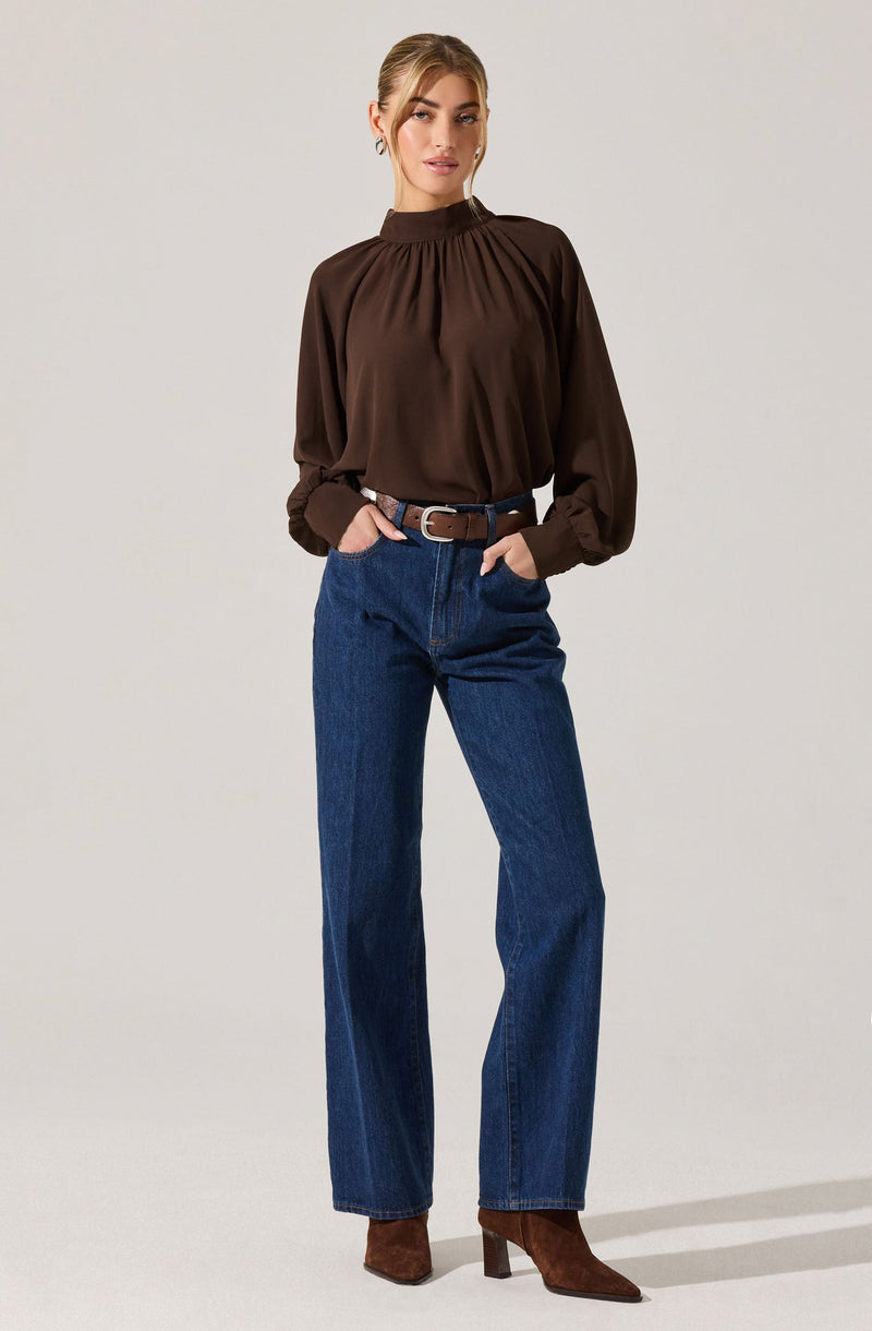 A woman is wearing a dark brown long-sleeve blouse with a high, gathered neckline and billowy sleeves that are cinched at the wrists. She pairs the blouse with high-waisted, straight-leg blue jeans secured with a brown belt, and completes the outfit with brown heeled ankle boots.