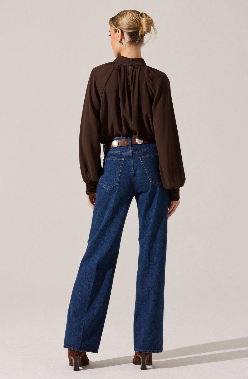 A woman is wearing a loose-fitting, long-sleeved chocolate brown blouse with gathered fabric at the neckline and cuffs. She pairs it with high-waisted, wide-leg blue jeans and brown heeled shoes. Her hair is styled in a neat bun.