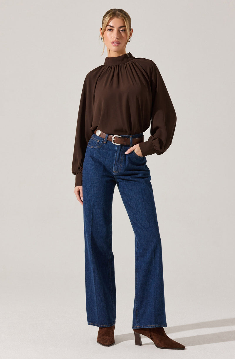 A woman is wearing a dark brown, long-sleeved blouse with a high, gathered neckline and puffed, loose sleeves that gather at the cuffs. She has paired it with high-waisted blue jeans, a brown belt, and brown ankle boots with a block heel.