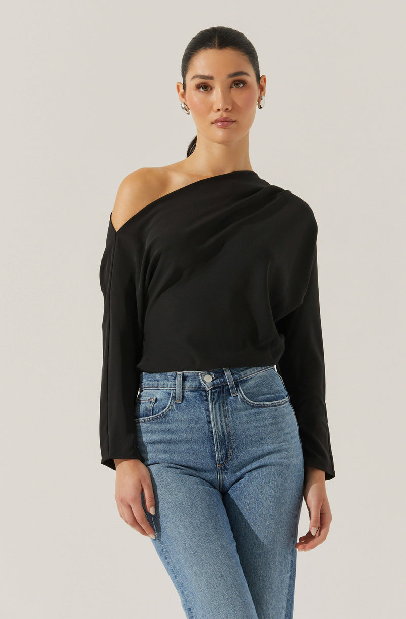 A woman is wearing a black satin blouse with a one-shoulder design, featuring long sleeves and a relaxed fit. She has paired it with high-waisted, light-wash blue jeans.
