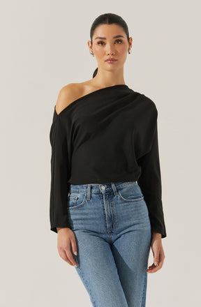 A woman is wearing a black satin blouse with a one-shoulder design, featuring long sleeves and a relaxed fit. She has paired it with high-waisted, light-wash blue jeans.