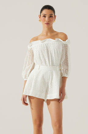 A woman is wearing an off-the-shoulder white eyelet lace top with elastic at the neckline and sleeves, and matching high-waisted shorts with a wide waistband and a flowy, relaxed fit. The top has puffed three-quarter length sleeves with a textured pattern.