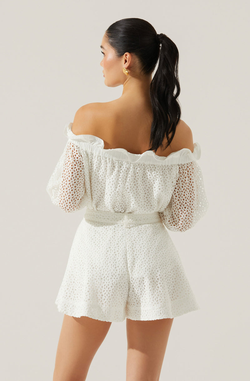 The woman is wearing a white off-the-shoulder top with puffed, long sleeves made of eyelet lace fabric, featuring a ruffled neckline. She is also wearing matching high-waisted shorts with a similar eyelet lace pattern, giving a coordinated and stylish look.