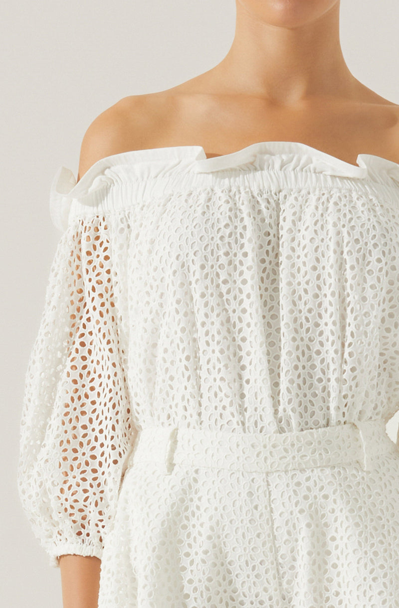 A woman is wearing a white, off-the-shoulder dress with a textured, eyelet pattern. The dress features elasticized ruffles along the neckline and long sleeves with elastic at the cuffs. The waistline is cinched with a matching fabric belt, creating a gathered effect.