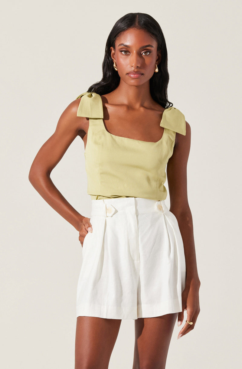 A woman is wearing a sleeveless, light yellow top with shoulder bows and wide straps. She pairs it with high-waisted, white pleated shorts that have belt loops.