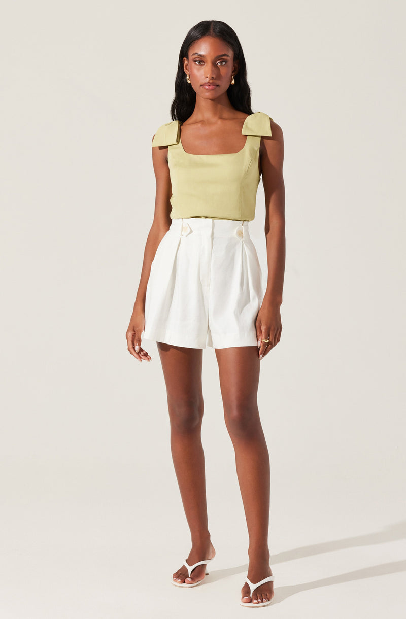 A woman is wearing a sleeveless, light yellow top with wide shoulder straps that have decorative bow details, paired with high-waisted, white tailored shorts with pleats and decorative buttons at the waist. She is also wearing white, open-toed high-heeled sandals.
