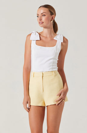 A woman is wearing a sleeveless white top with large bow details on the shoulder straps and a square neckline. She pairs the top with high-waisted, light yellow textured shorts.