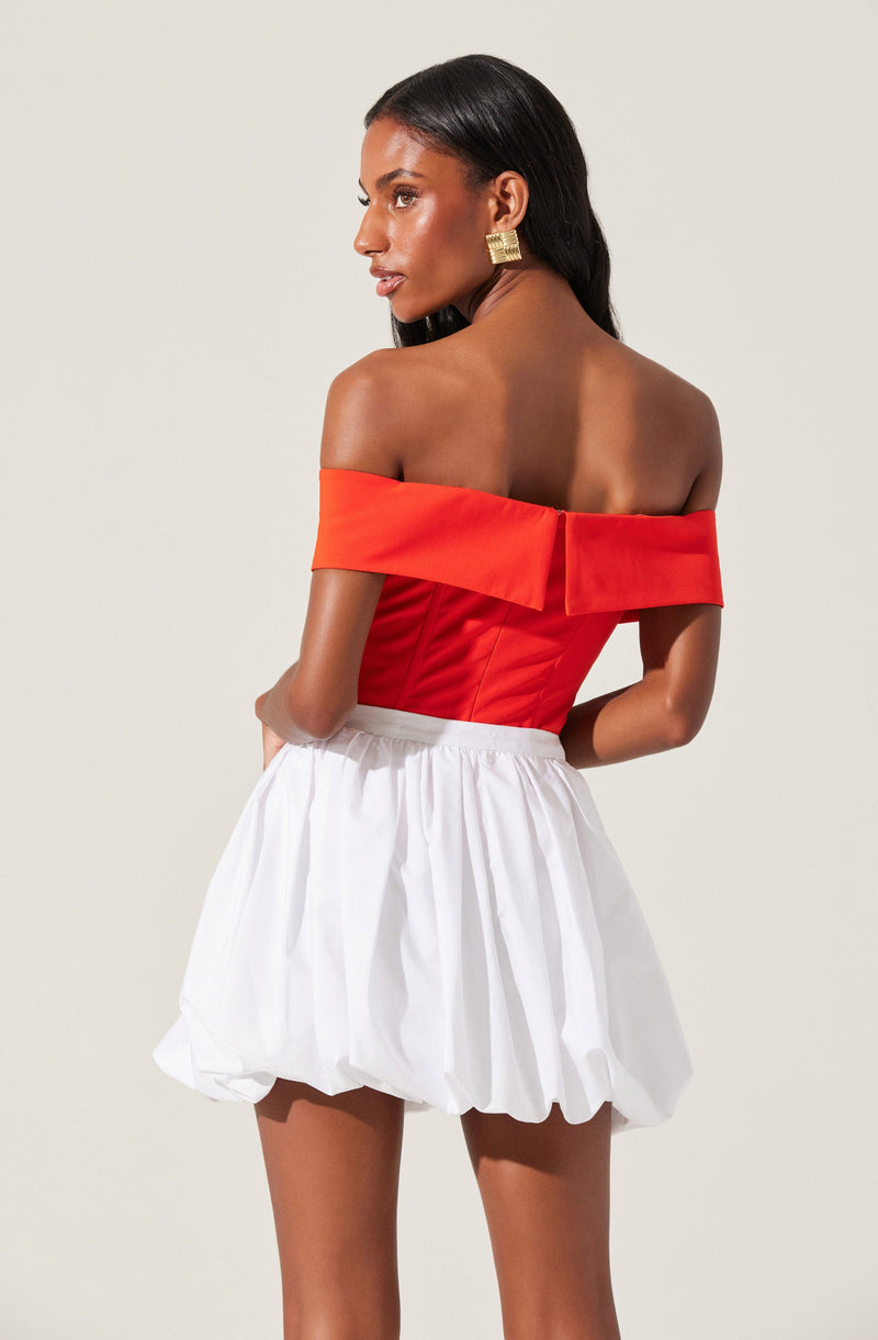 A woman is wearing an off-the-shoulder red top with a wide folded neckline and a fitted bodice, paired with a white, billowy, knee-length skirt that has gathered fabric creating a textured, voluminous appearance.