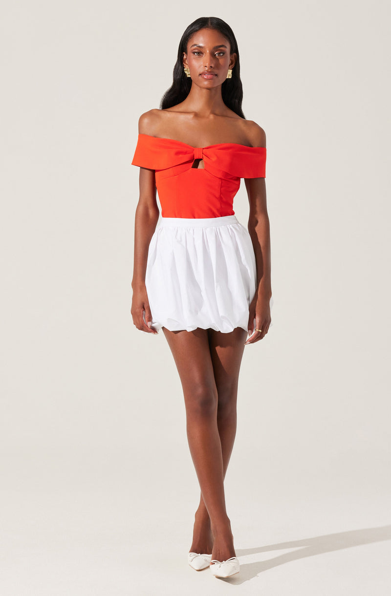 A woman is wearing an off-the-shoulder, bright red top with a knot and cut-out detail at the chest. She pairs it with a high-waisted white skirt that is gathered and slightly puffy, ending above the knees. She completes her outfit with white pointed-toe heels.