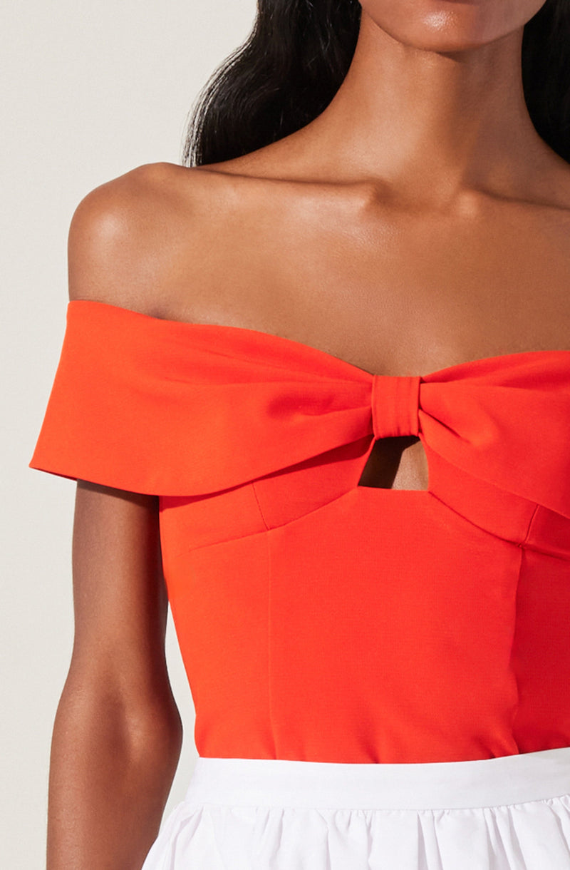 A woman is wearing an off-the-shoulder, bright red top with a large bow detail at the chest that has a central knot and a small keyhole opening below it. The top appears to be made of a smooth, structured fabric. She is also wearing a white skirt with a gathered waistband.