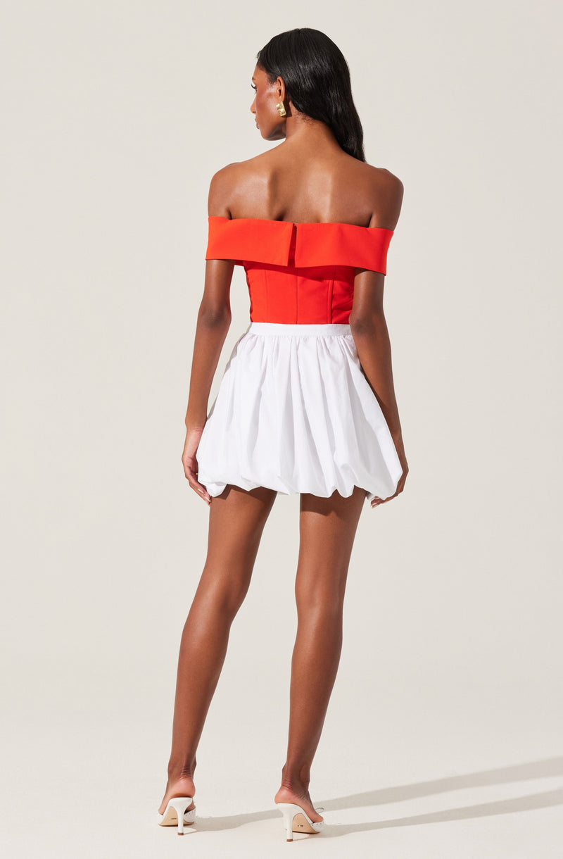 A woman is wearing an off-the-shoulder red top with a wide folded neckline and a fitted bodice, paired with a high-waisted white puffed skirt that has a gathered, balloon-like shape. She is also wearing white high-heeled sandals.