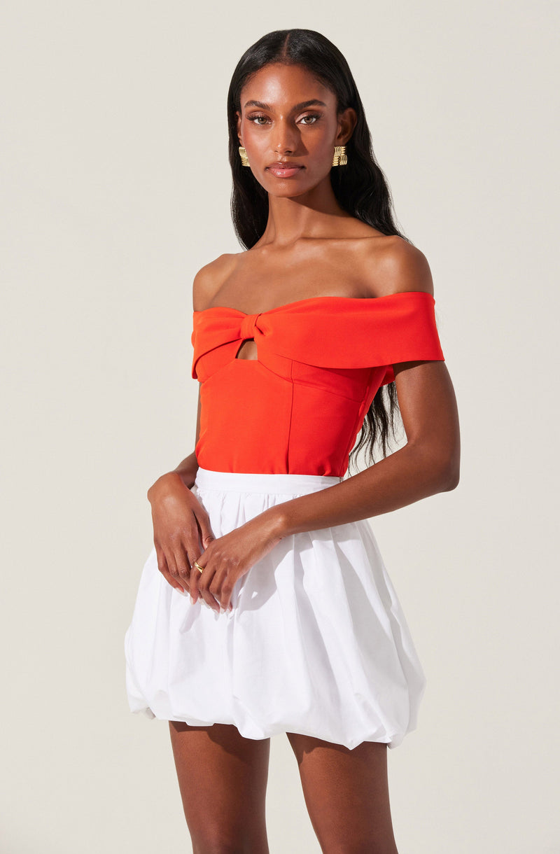 A woman is wearing an off-the-shoulder, bright orange top with a knot detail at the front and a small cut-out below the knot. She pairs it with a white, voluminous, bubble-style skirt that has an elastic waistband. She has long, dark hair and is accessorized with gold earrings and a ring.
