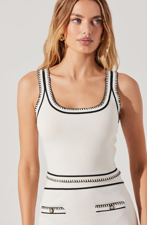 A woman is wearing a sleeveless, fitted, cream-colored top with bold black trim details around the neckline, armholes, waist, and pockets. The top has a scoop neckline and decorative stitching along the edges, creating a contrasting pattern. She has her hair styled in loose waves and is accessorized with gold earrings.