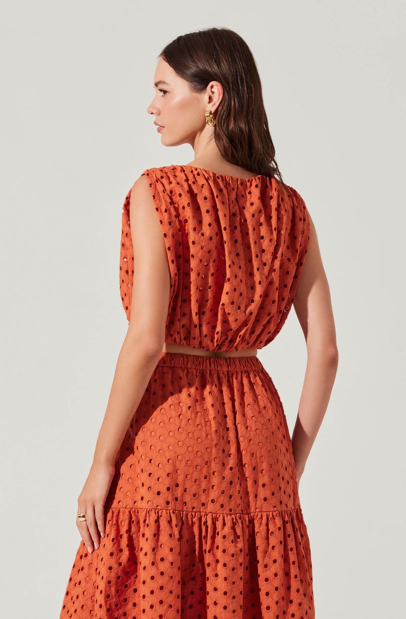 A woman is wearing a two-piece outfit in a burnt orange color with a textured, semi-sheer fabric featuring black polka dots. The top is sleeveless with gathered fabric at the neckline and a loose fit, cropped just above the waist. The matching skirt has an elastic waistband, a similar polka dot pattern, and a tiered, ruffled design that extends to a midi length. The overall look is stylish and summery.