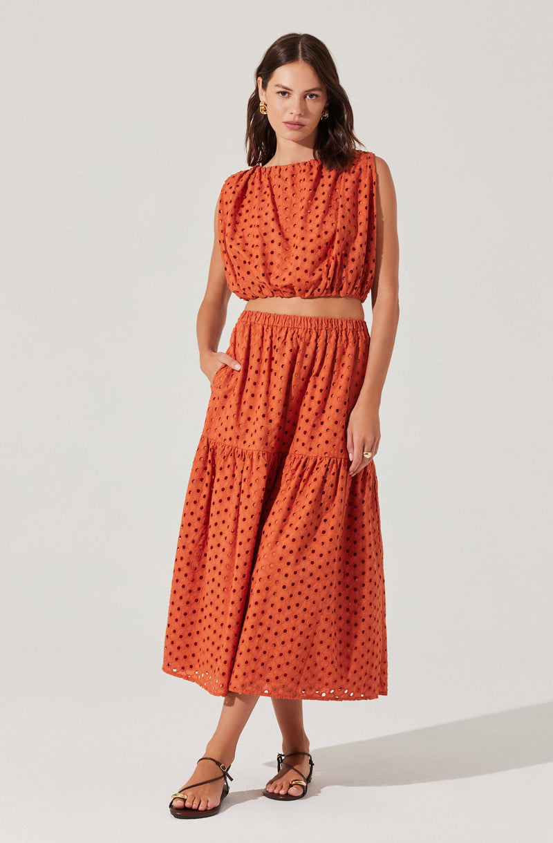 A woman is wearing a sleeveless, cropped top and a matching high-waisted, wide-leg skirt in a bright orange fabric with a black polka dot pattern. The top has a loose fit with gathered fabric at the neckline and armholes. The skirt is flowy, reaching mid-calf length, with a tiered design. The outfit is paired with black strappy sandals and gold hoop earrings.