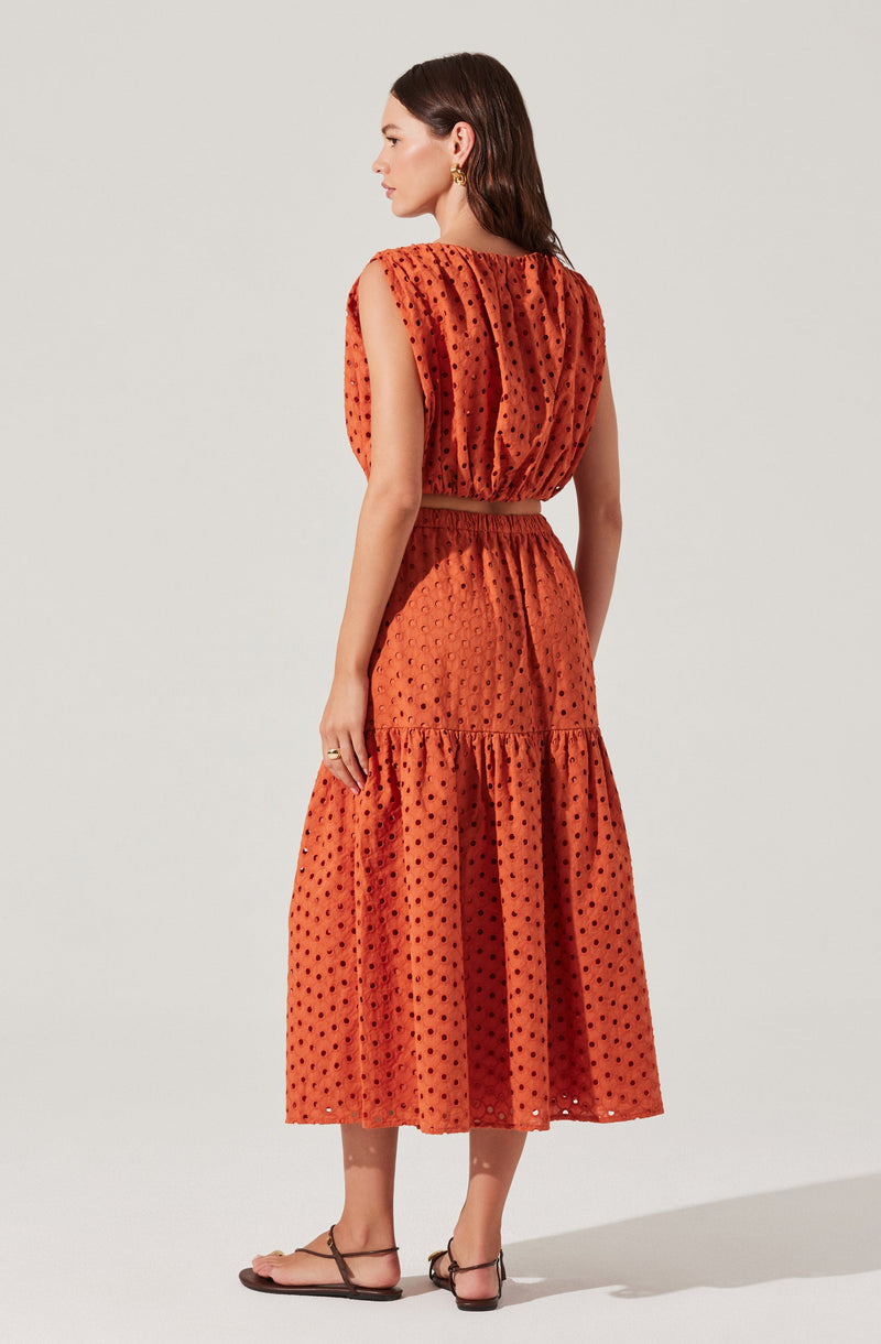 A woman is wearing a sleeveless, burnt orange dress with a dotted pattern. The top of the dress is loose-fitting with a gathered neckline. The waistline is elasticized, and the skirt is tiered, with the lower tier being fuller and longer. She is paired the outfit with flat, strappy sandals.